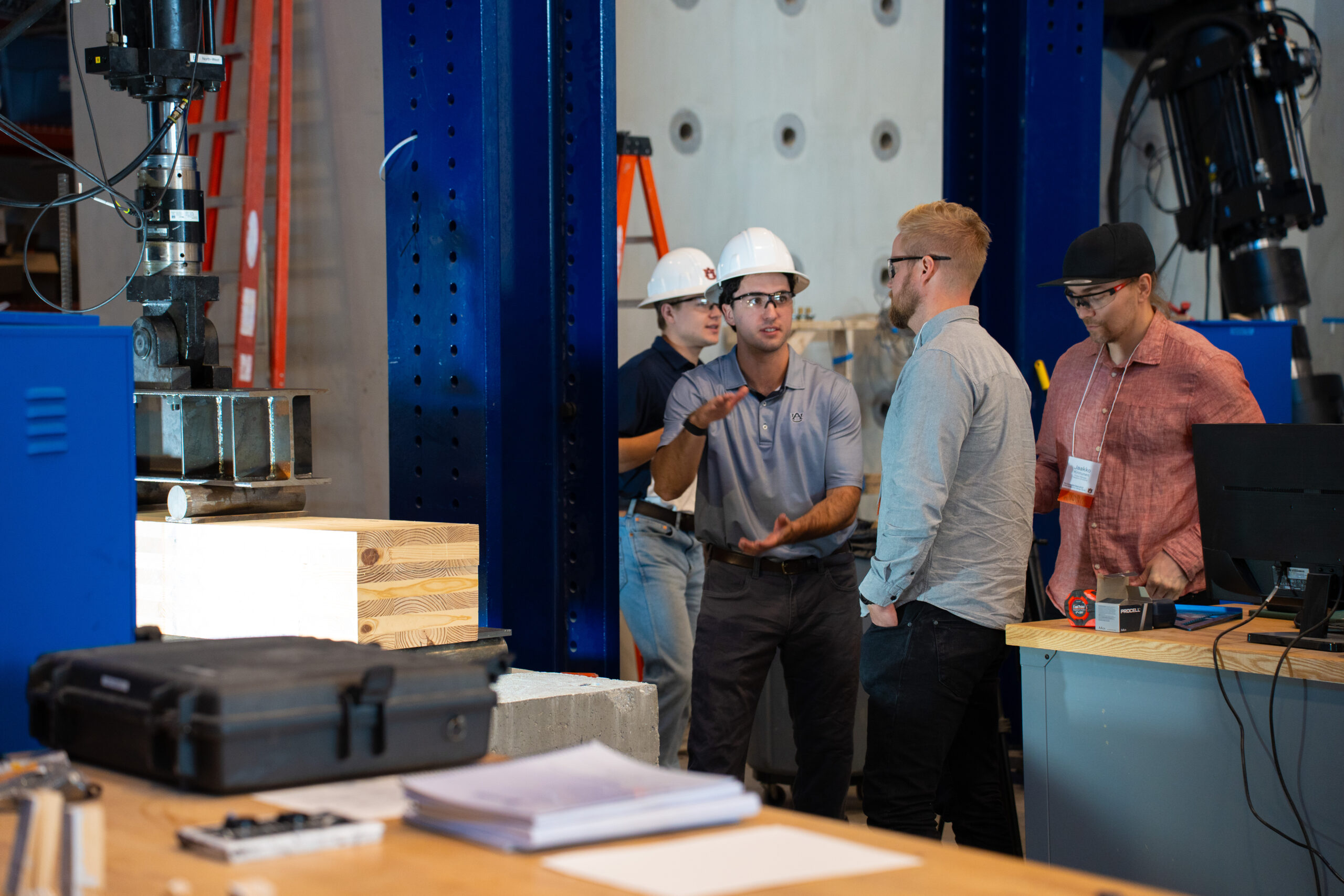 Men in hard hats demonstrate the strength of the laminated wood during a laboratory tour.